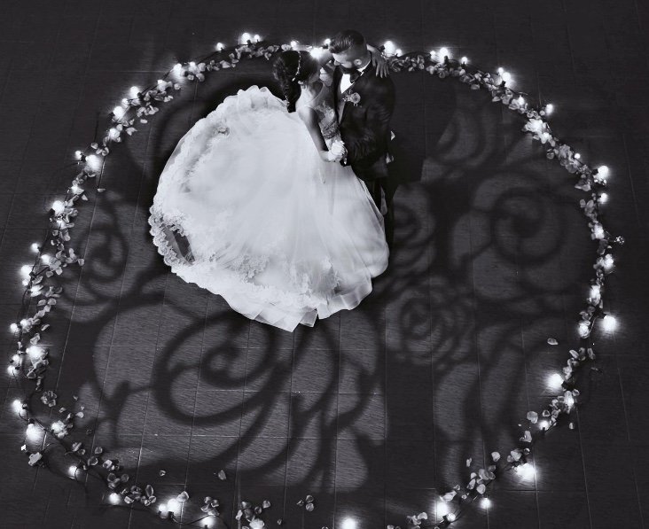 A couple dances tenderly in a circular arrangement of rose petals and soft lights, casting romantic shadows on the floor in black and white.