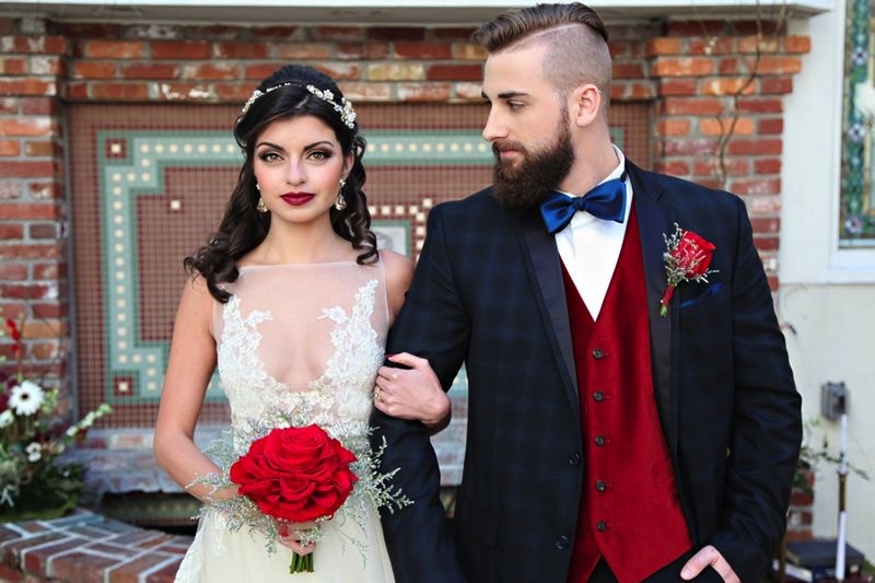 Bride in an elegant lace gown holds a bouquet of red roses, standing beside a stylish groom in a dark suit. They appear ready for a wedding ceremony.
