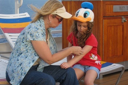 A woman helps a young girl, who wears a large Donald Duck hat, on a cruise ship deck. They are seated near colorful lounge chairs.