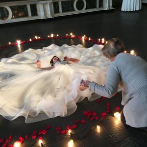 A bride in a flowing white gown lies on the floor surrounded by rose petals and soft lights, while a woman adjusts her dress.
