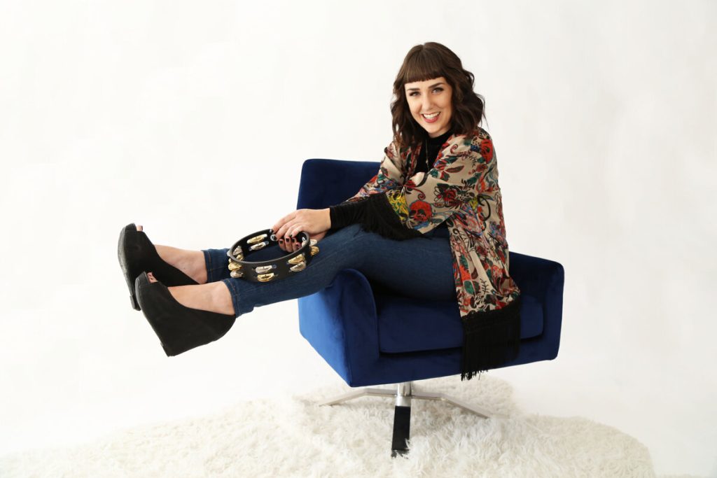Smiling woman in a floral shawl sits on a blue chair, holding a tambourine, showcasing a stylish and joyful music-themed vibe.