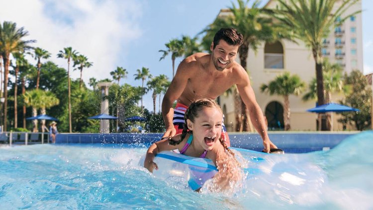 Father and daughter enjoy a splash-filled moment at a resort pool, surrounded by palm trees and sunny skies. Perfect for family fun and relaxation.