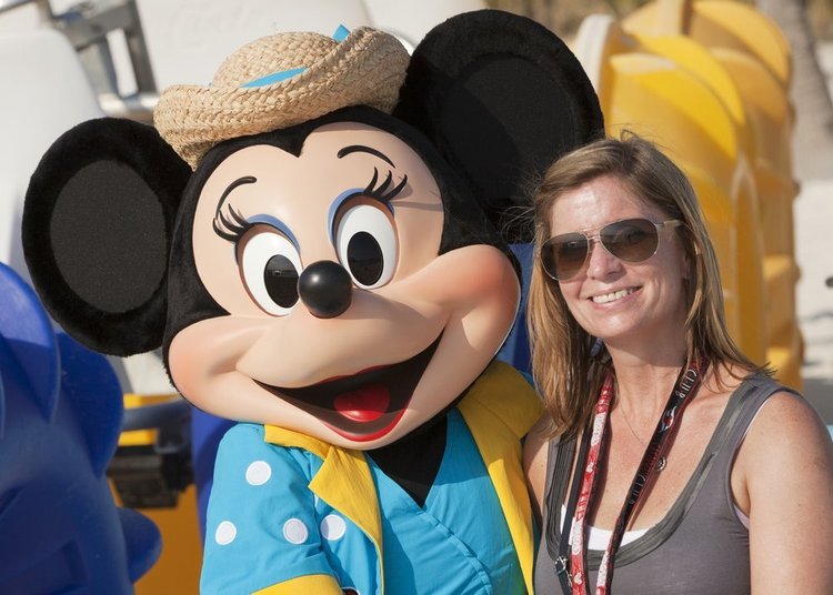 Minnie Mouse, dressed in a colorful outfit and straw hat, poses happily with a woman in sunglasses at a beach setting.