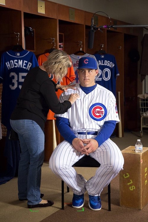 A baseball player in a Chicago Cubs uniform sits on a stool in a locker room, while a woman adjusts his jersey for a photo shoot.