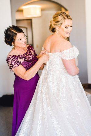A bride in a stunning lace gown smiles as her mother fastens her dress, capturing an intimate moment of preparation for the wedding day.