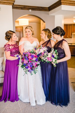 A bride in a white gown smiles with her three bridesmaids, dressed in purple tones, as they hold vibrant bouquets featuring pink and purple flowers.
