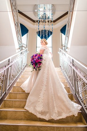 Bridal portrait of a woman in a lace wedding gown ascending elegant stairs, holding a vibrant bouquet, with chandeliers and blue accents in the background.