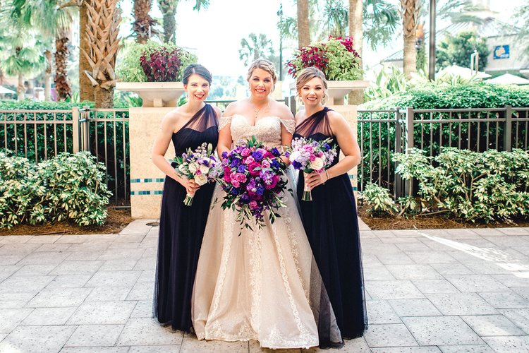 Bridal party posing outdoors, featuring the bride in an embellished gown and two bridesmaids in navy dresses, each holding vibrant flower bouquets.