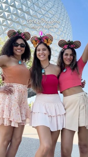 Three friends pose together in front of a large geodesic dome at a theme park, wearing playful outfits and floral headbands.