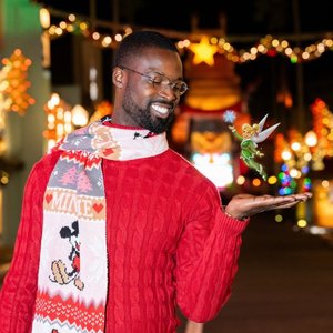 A man in a red sweater and festive scarf smiles while gently holding Tinker Bell on his hand, surrounded by colorful holiday lights.