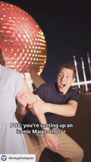 A joyful man gestures excitedly while assisting in setting up a Magic Shot photoshoot at night, with a glowing backdrop of an iconic structure.