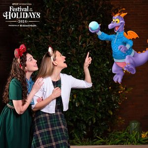 Two women, dressed festively, gaze excitedly at a purple dragon character in a sweater, who holds a snowball, at the Epcot Festival of Holidays 2024.