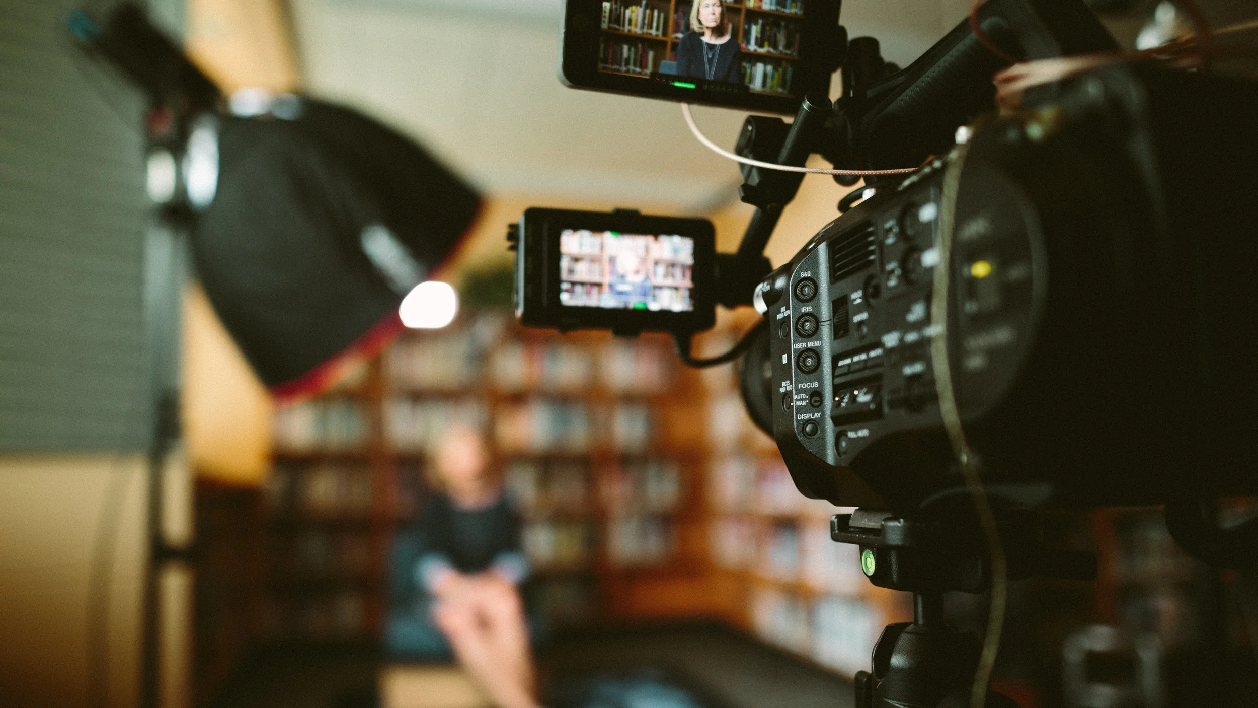 Camera equipment is set up in a library for a video interview. A person is framed on a monitor, highlighting a professional filming environment.