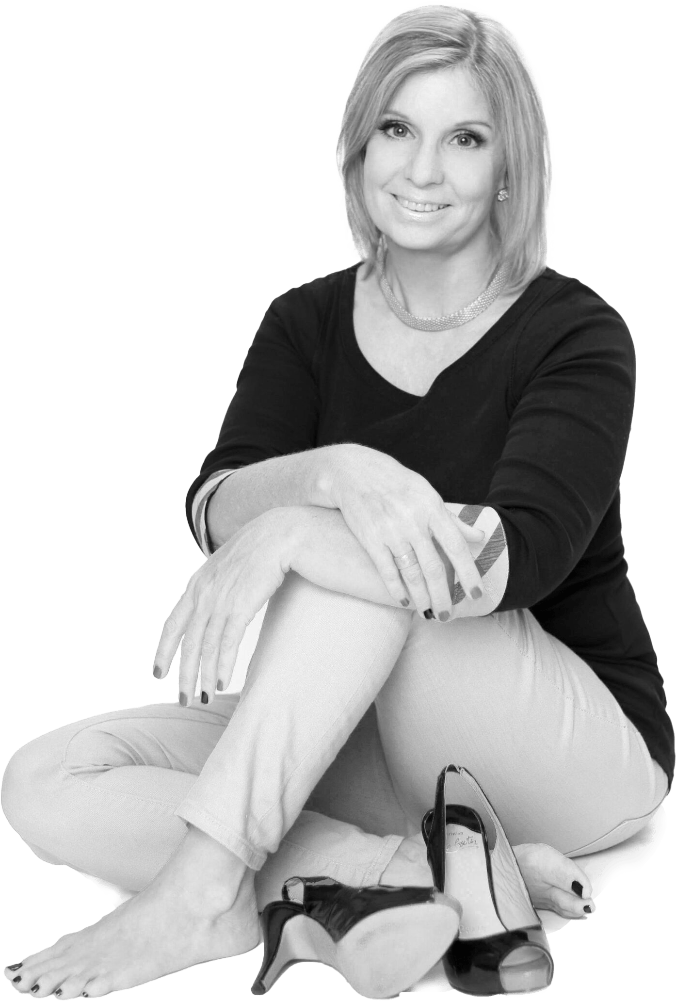 A smiling woman sits cross-legged, barefoot, beside a pair of high-heeled shoes, showcasing a relaxed and fashionable vibe.