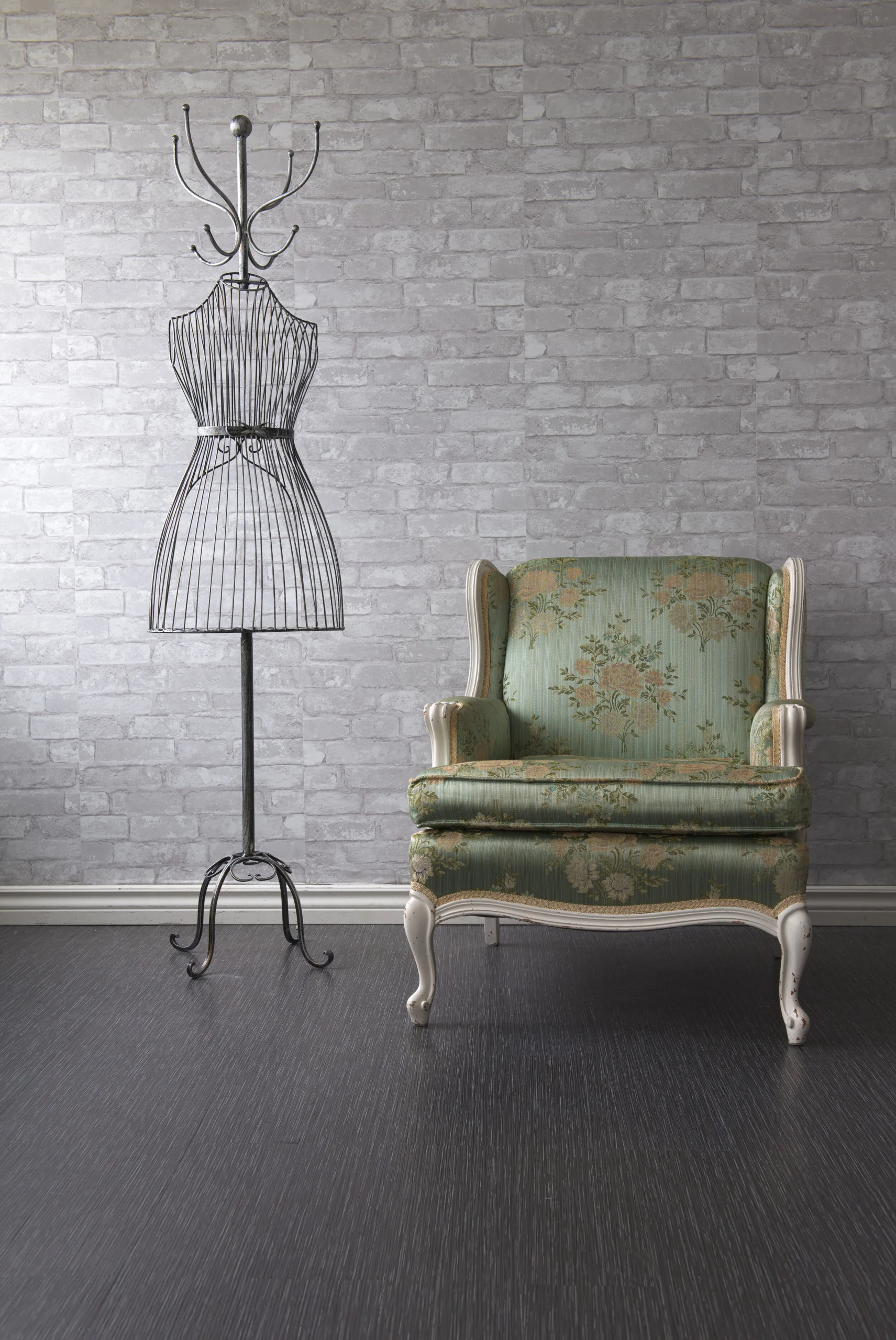 Wire dress form stands beside a vintage green upholstered chair against a gray brick wall, showcasing a stylish, eclectic interior design.