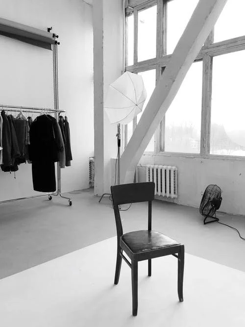 Minimalist studio setup featuring a wooden chair, clothing rack, lighting umbrella, and natural light through large windows, ideal for a photoshoot.
