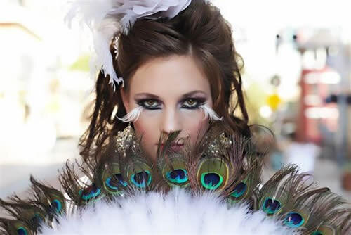 A model with dramatic makeup and intricate peacock feather fan poses confidently, showcasing vibrant colors and artistic expression, likely for a fashion shoot.